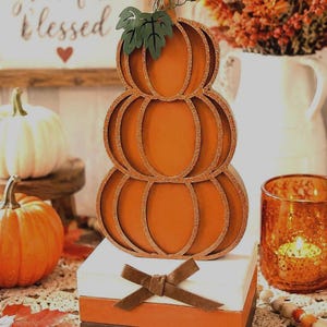 May include: A stacked orange pumpkin decoration with a green metal leaf accent, resting on a base of three rectangular blocks in white, orange, and dark brown, featuring a brown fabric bow tied around the top white block.