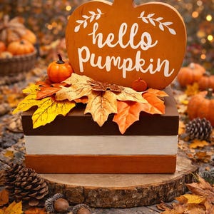 May include: A wooden pumpkin-shaped sign with the words "hello Pumpkin" in white script. The sign sits atop a stack of three wooden blocks, decorated with fall leaves and small pumpkins. The scene is set on a wooden surface with pine cones and acorns.