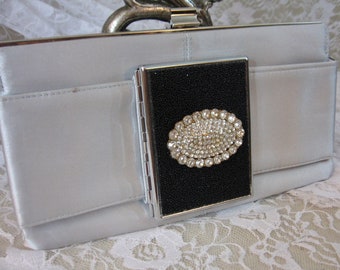 May include: A silver satin clutch with a black and silver hinged compartment. The compartment features a large oval-shaped rhinestone brooch.