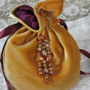 May include: A golden velvet drawstring pouch with a deep purple interior and matching ribbon ties. The pouch is adorned with a beaded tassel featuring amber, gold, and red beads. The background is a white lace fabric.