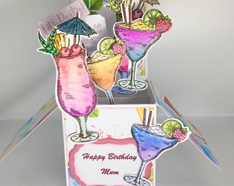 May include: A pop-up card with a white background and a pink, red, and white message that says "Happy Birthday Mum". The card features a pop-up design with four colorful cocktails with fruit and umbrellas.
