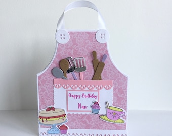 May include: Pink and white apron-shaped card with a pocket and a "Happy Birthday Nan" message. The card features a cake, cupcake, teacup, and kitchen utensils.
