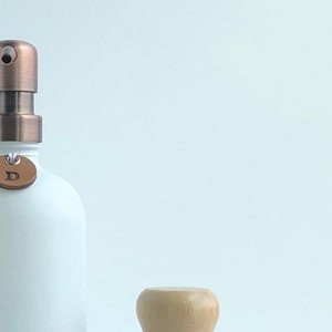 Może przedstawiać: Biały ceramiczny dozownik do mydła z pompką miedzianą i brązową sk&oacute;rzaną etykietą. Dozownik stoi na białym marmurowym blacie obok białej ceramicznej miski z drewnianym pędzlem.