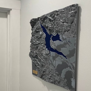 May include: A gray and blue 3D topographical map of a mountainous region, mounted on a wall. The map features a blue lake and detailed relief, showcasing the terrain's elevation. The artwork is displayed near a white door.