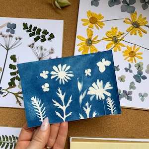 May include: A cyan blue card with white silhouettes of flowers and leaves. The card is being held by a hand with a ring on the finger.