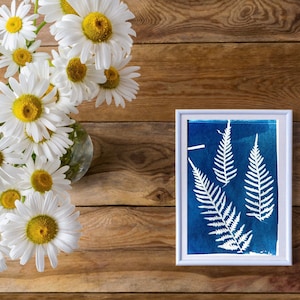 May include: A white frame with a blue background and a white fern silhouette inside. A bouquet of white daisies with yellow centers is in a glass vase on a wooden table.