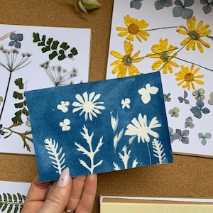 May include: A blue card with white pressed flowers and leaves. The card is held in a hand.
