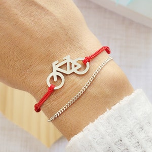 May include: A red string bracelet with a silver bicycle charm. The bracelet is on a person's wrist.