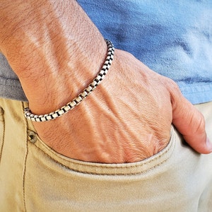 May include: A silver chain bracelet with a box chain design. The bracelet is worn on a person's wrist.