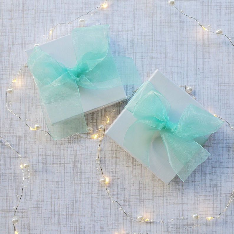 May include: Two white gift boxes with light blue sheer ribbon bows. The boxes are on a white textured background with a string of white lights.