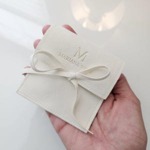 May include: A white velvet pouch with a gold ribbon tied in a bow. The pouch has the letter "M" and the text "Mariana Vita" embossed in gold.