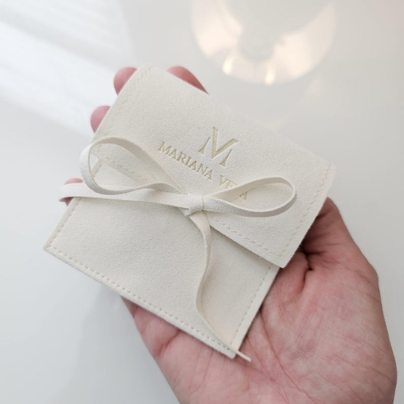 May include: A white velvet pouch with a gold ribbon tied in a bow. The pouch has the letter "M" and the text "Mariana Vita" embossed in gold.