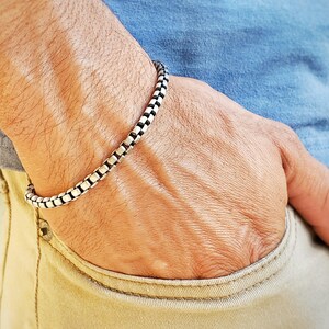 May include: A silver chain bracelet with a box chain design. The bracelet is worn on a person's wrist.