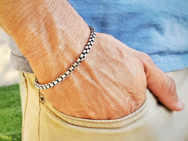 May include: A silver chain bracelet with a box chain design. The bracelet is worn on a person's wrist.