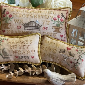 May include: Three embroidered pillow covers with a yellow rope trim. The first pillow cover features the alphabet and a bench. The second pillow cover has the text "Always be my heart 1908". The third pillow cover has two birds on a pedestal and a vine with red berries.