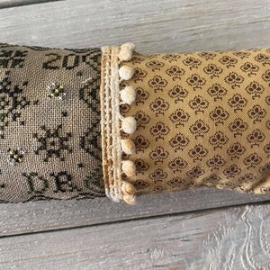 Op de afbeelding: Een beige en bruin kussen met een kruissteekontwerp aan de ene kant en een bruin bloemenpatroon aan de andere kant. Het kussen heeft een witte pomponrand.