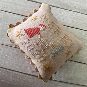 May include: A beige pillow with a scalloped edge features a cross-stitch design of a red bird, trees, and stars. The text "Merry Christmas" is stitched in brown thread.