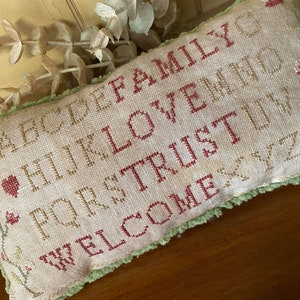 May include: A beige cushion with green fringe, embroidered with the words "ABCDE FAMILY HIJK LOVE FGHI TRUSTUVWXYZ WELCOME" in red and brown thread.