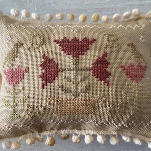 May include: A small, beige pillow with a white pom-pom trim. The pillow is embroidered with a floral design featuring red tulips and green leaves.