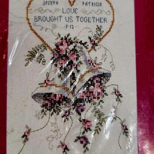 May include: Cross-stitch wedding sampler with a heart-shaped border, the names "Joseph" and "Patricia", and the phrase "Love Brought Us Together." Below the heart are two bells surrounded by pink and purple flowers.