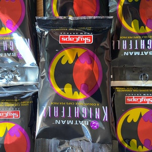 Może przedstawiać: Zbliżenie na zamknięte opakowanie kolekcjonerskich kart SkyCaps Batman Knightfall. Opakowanie jest czarne z fioletowym i żółtym logo Batmana. Tekst na opakowaniu brzmi "Batman Knightfall 6 kolekcjonerskich kart w opakowaniu".