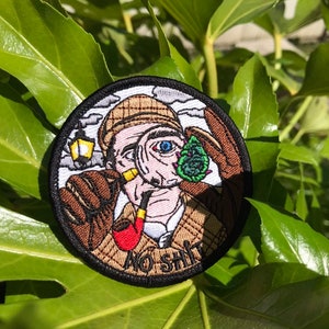 May include: A round embroidered patch with a cartoon image of a man wearing a hat and a magnifying glass. The man has a red pipe in his mouth and is looking at a green and blue object. The text "NO SHIT" is below the image.