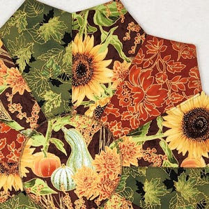 May include: A circular fabric table mat featuring a patchwork design of autumn-themed patterns. The fabric displays sunflowers, orange pumpkins, green gourds, and maple leaves in shades of brown, dark green, orange, and yellow, arranged in a starburst.
