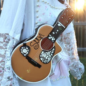Puede incluir: Un bolso marrón con forma de guitarra con detalles florales blancos y el nombre "SUGA" en escritura negra. El bolso incluye un charm en forma de púa con un diseño floral. Tiene una correa marrón y ribete blanco.