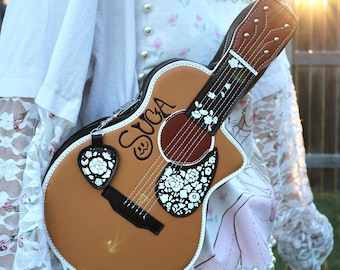 May include: A brown guitar-shaped handbag with white floral accents and the name "SUGA" in black script. The bag features a guitar pick-shaped charm with a floral design. The bag has a brown strap and a white trim.