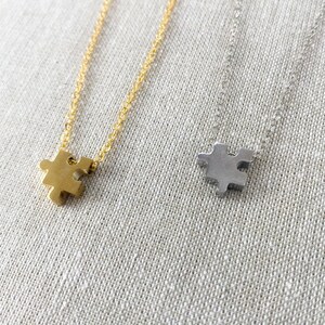 May include: Two necklaces featuring small puzzle piece pendants. One pendant is gold-toned and hangs from a matching gold-toned chain, while the other is silver-toned and hangs from a matching silver-toned chain. Both chains are delicate and link-style.