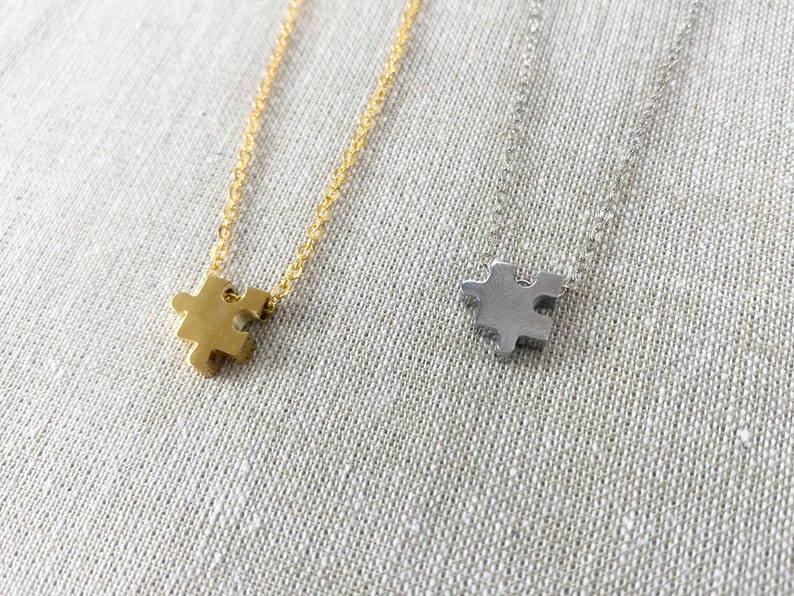 May include: Two necklaces featuring small puzzle piece pendants. One pendant is gold-toned and hangs from a matching gold-toned chain, while the other is silver-toned and hangs from a matching silver-toned chain. Both chains are delicate and link-style.