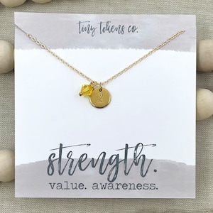 May include: Gold necklace with a small round charm that says "Strength" and a yellow crystal bead. The necklace is on a white card with the text "tiny tokens co." and "value. awareness."