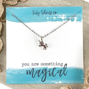 May include: A silver necklace with a small unicorn charm. The necklace is displayed on a blue and white card with the text "tiny tokens co." and "you are something magical".