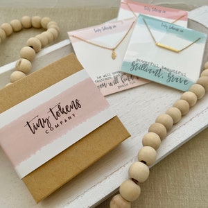 May include: A brown cardboard gift box with a pink label reading tiny tokens company, accompanied by two gold necklaces on display cards. One card features a feather charm and Psalm 91:4 text, while the other shows a bar pendant and the text powerful, beautiful, brilliant, brave.