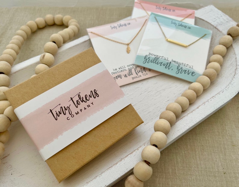 May include: A brown cardboard gift box with a pink label reading tiny tokens company, accompanied by two gold necklaces on display cards. One card features a feather charm and Psalm 91:4 text, while the other shows a bar pendant and the text powerful, beautiful, brilliant, brave.
