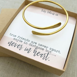 May include: A gold-colored open cuff bracelet with leaf-like ends rests on a card inside a small box. The card has the text "true friends are never apart, maybe in distance but never in heart." The box is light brown.