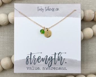 May include: A gold-colored necklace with a small green crystal bead and a round charm with a ribbon symbol. The necklace is displayed on a white card with the words "strength. value. awareness." printed in black.