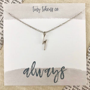 May include: A silver necklace with a lightning bolt charm and the word "always" written in cursive on a white card with a gray background.
