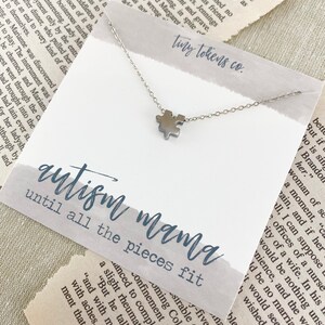 May include: A silver-toned puzzle piece charm necklace on a white display card. The card features the text "tiny tokens co." at the top and "autism mama until all the pieces fit" in blue script and block lettering across the center.