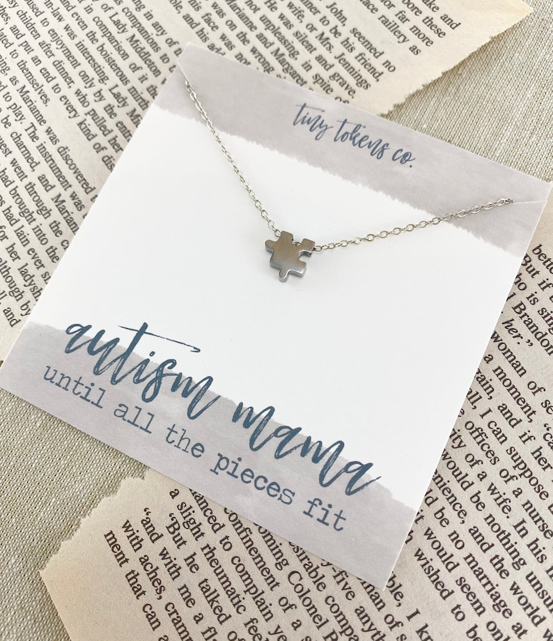 May include: A silver-toned puzzle piece charm necklace on a white display card. The card features the text "tiny tokens co." at the top and "autism mama until all the pieces fit" in blue script and block lettering across the center.