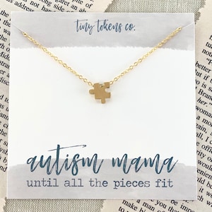 May include: A gold-toned necklace featuring a small puzzle piece pendant on a delicate chain, displayed on a white card with the text tiny tokens co., autism mama, and until all the pieces fit printed in blue script and block lettering.