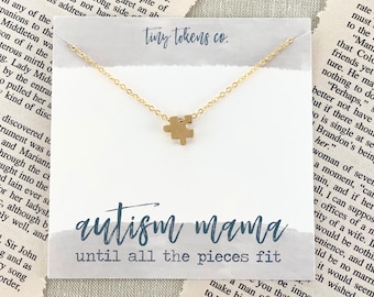 May include: A gold-toned necklace featuring a small puzzle piece pendant on a delicate chain, displayed on a white card with the text tiny tokens co., autism mama, and until all the pieces fit printed in blue script and block lettering.
