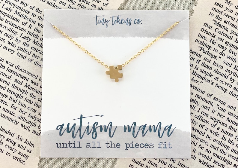 May include: A gold-toned necklace featuring a small puzzle piece pendant on a delicate chain, displayed on a white card with the text tiny tokens co., autism mama, and until all the pieces fit printed in blue script and block lettering.