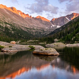May include: A mountain lake reflecting the orange and pink hues of a sunset sky. The lake is surrounded by a forest of evergreen trees and rocky mountains.