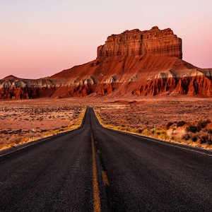 May include: A long, straight asphalt road leads towards a large, red rock formation in a desert landscape. The sky is a soft pink and purple.