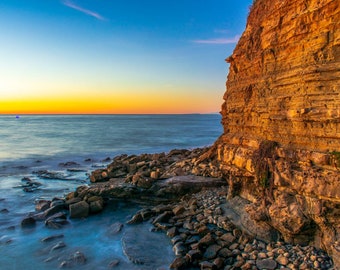 May include: A rocky coastline with a cliff face at sunset. The water is a deep blue and the sky is a vibrant orange and pink. The cliff face is a warm brown color with layers of rock.
