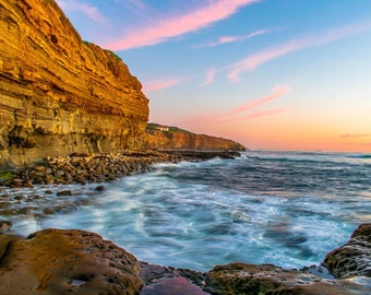 May include: Coastal landscape featuring a layered sandstone cliff illuminated by the golden light of the setting sun. The ocean waves crash against the rocky shore, under a sky painted with pink and blue hues. A serene and natural scene.
