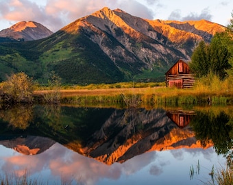 May include: A rustic wooden cabin with a red door sits in front of a lake reflecting the golden light of the setting sun behind a mountain range.
