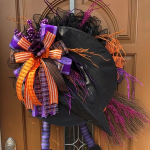 May include: A black witch hat with a purple and orange bow, striped legs, and black shoes. The hat is decorated with black mesh and purple and orange sprigs. The witch is hanging on a wooden door.