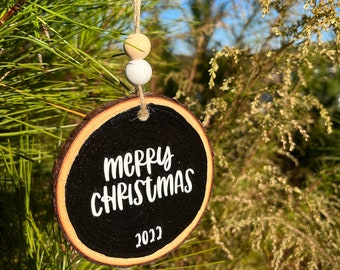 Może przedstawiać: Drewniana ozdoba w kształcie plastra, w kolorze czarnym, z napisem "merry christmas 2022" w białym kolorze. Ozdoba jest zawieszona na gałęzi sosny za pomocą brązowego sznurka jutowego i trzech drewnianych koralików.